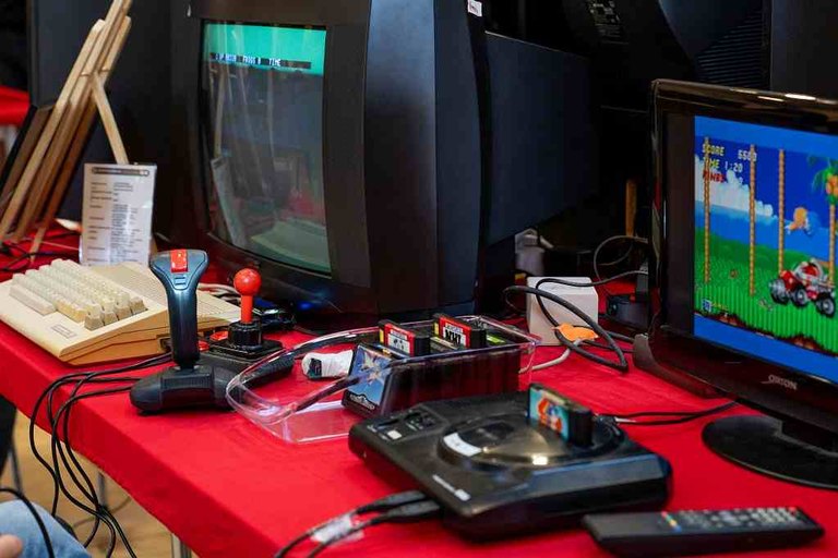 Retro-Spielkonsolen und ein Heimcomputer auf einem roten Tisch beim Gangelt Games Festival, auf den Röhrenfernsehern dahinter laufen Spiele
