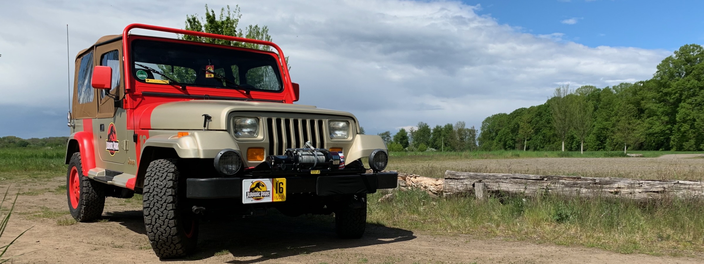 Frontansicht eines „Jurassic Park“-Jeeps auf einem Feldweg, dahinter eine Wiese und ein Waldrand